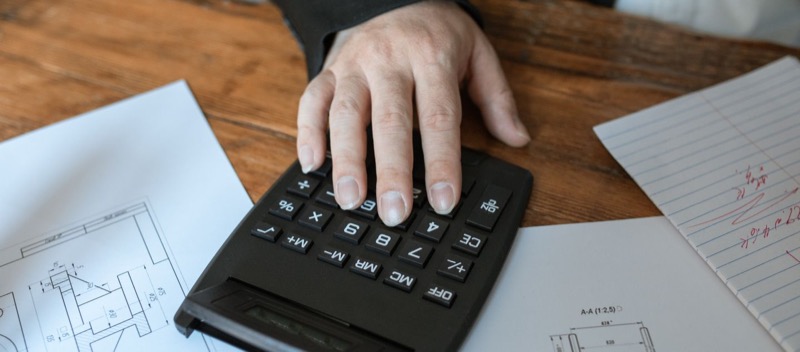 Une main pose les doigts sur les touches d'une calculatrice, sur un bureau en bois couvert de documents.