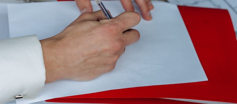 Une main écrit au stylo sur un document glissé dans une chemise rouge.