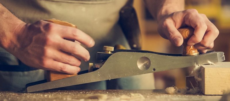 Les mains d'un menuisier poussent un rabot sur une planche, dans son atelier.