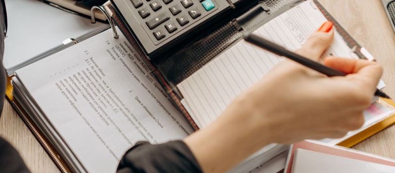Des mains utilisent une calculatrice posée sur un cahier de comptes.